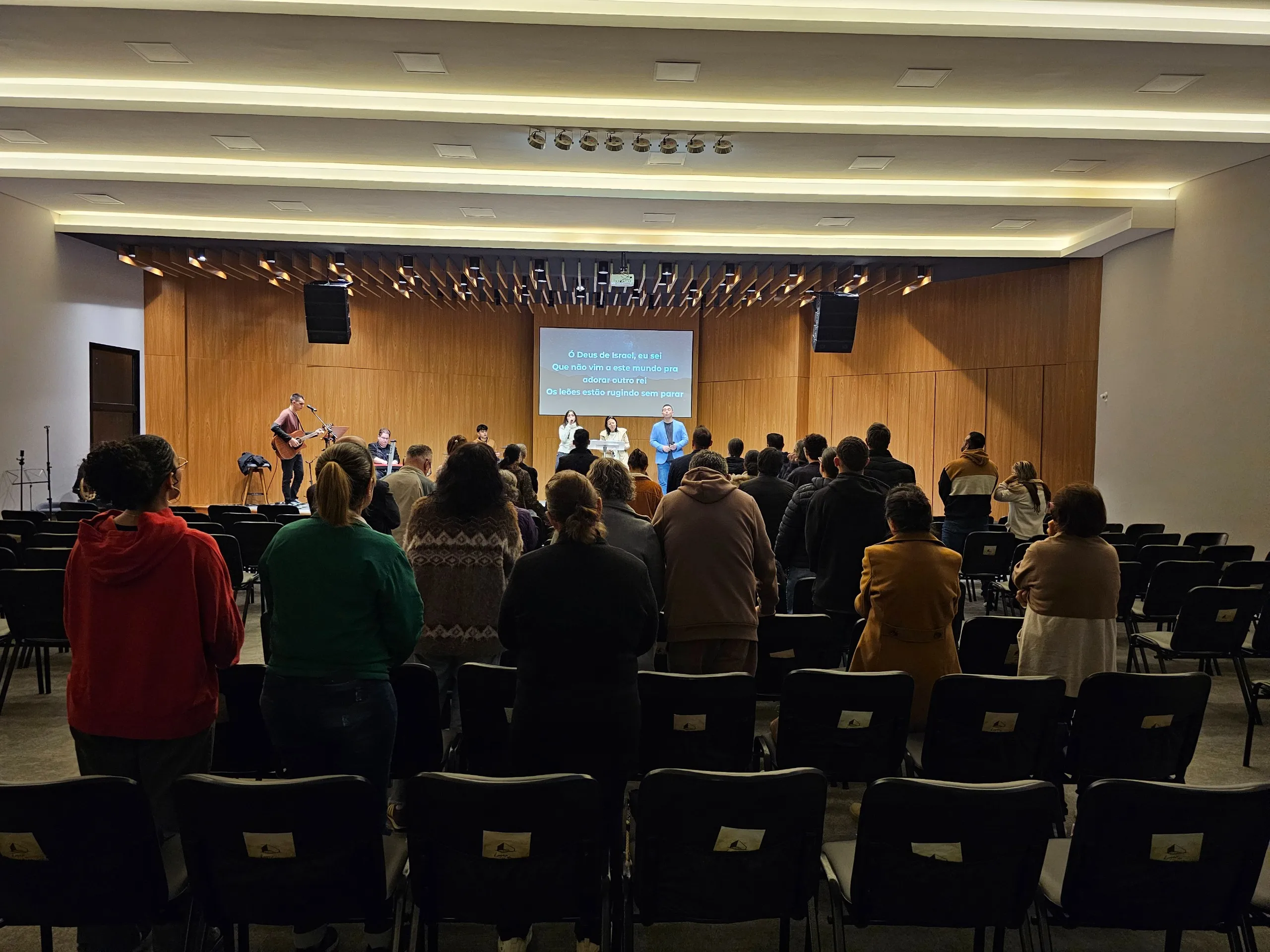 A congregação reunida em pé durante o culto, com a banda de louvor ao fundo